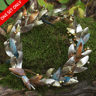 Circular metal wreath with overlapping leaf-shaped elements in various metallic finishes, adorned with small beads and resting on moss.