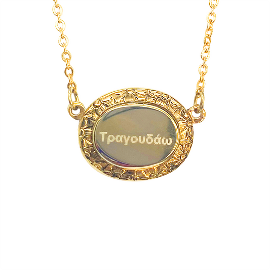 Gold oval pendant necklace with "Τραγουδάω" engraved in the center, surrounded by a floral border.