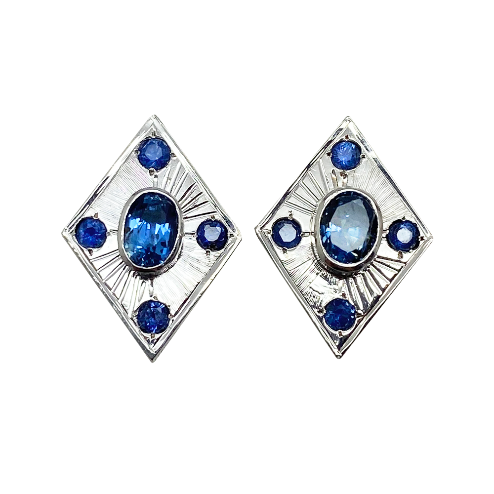 Pair of Art Deco-inspired earrings featuring a central oval blue sapphire surrounded by four smaller round blue sapphires. The earrings have a geometric, diamond-shaped design with intricate metalwork detailing.