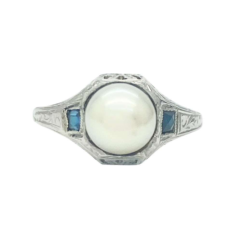 Silver ring featuring a large round pearl surrounded by a square-cut sapphire on each side.