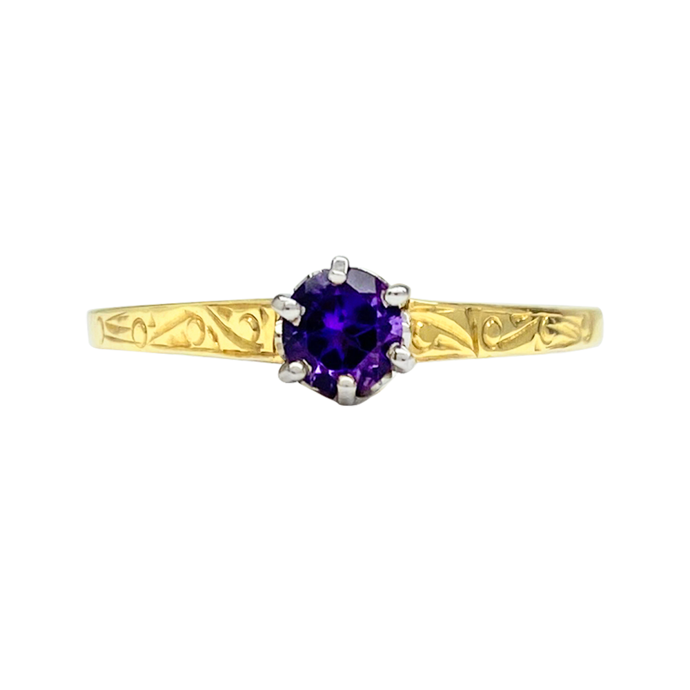 Gold ring with a round sapphire set in a four-prong setting. The band is engraved with intricate scrollwork.