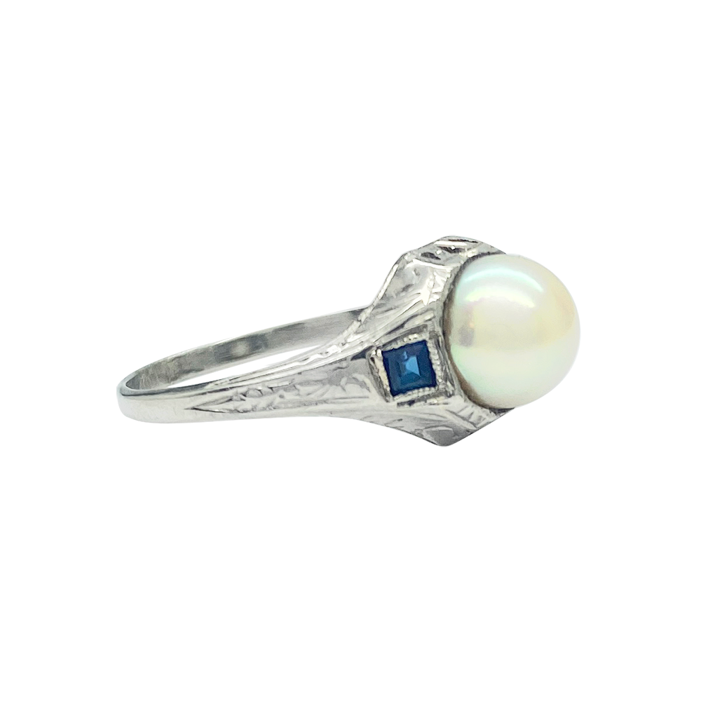 Silver ring featuring a large round pearl surrounded by a square-cut sapphire on each side.