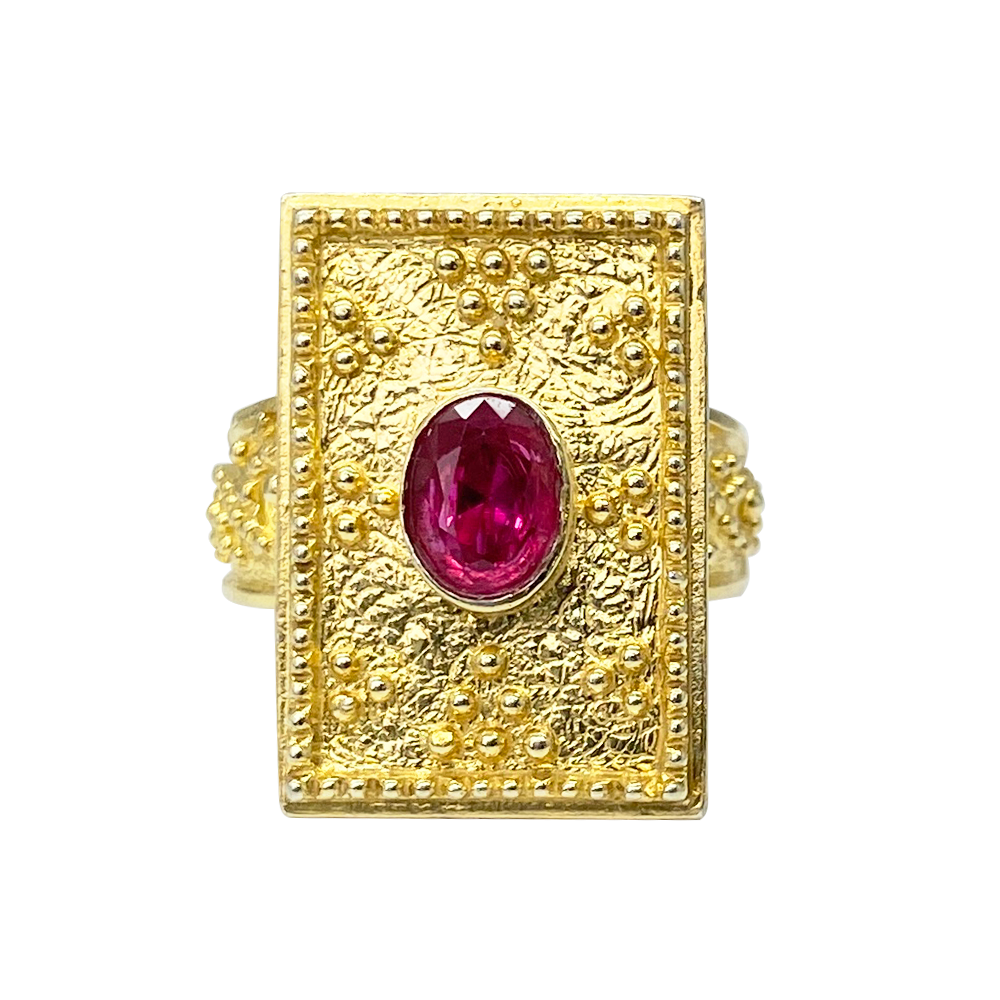 Gold ring with a square setting featuring an oval shaped ruby center stone and intricate textured details, on a white background.